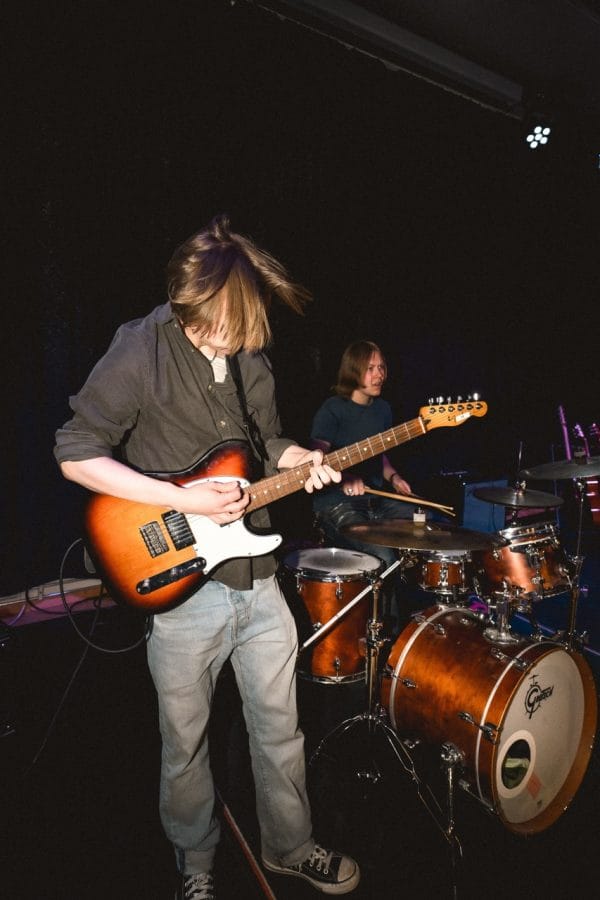 Kille spelar elgitarr. I bakgrunden sitter en kille och spelar trummor.